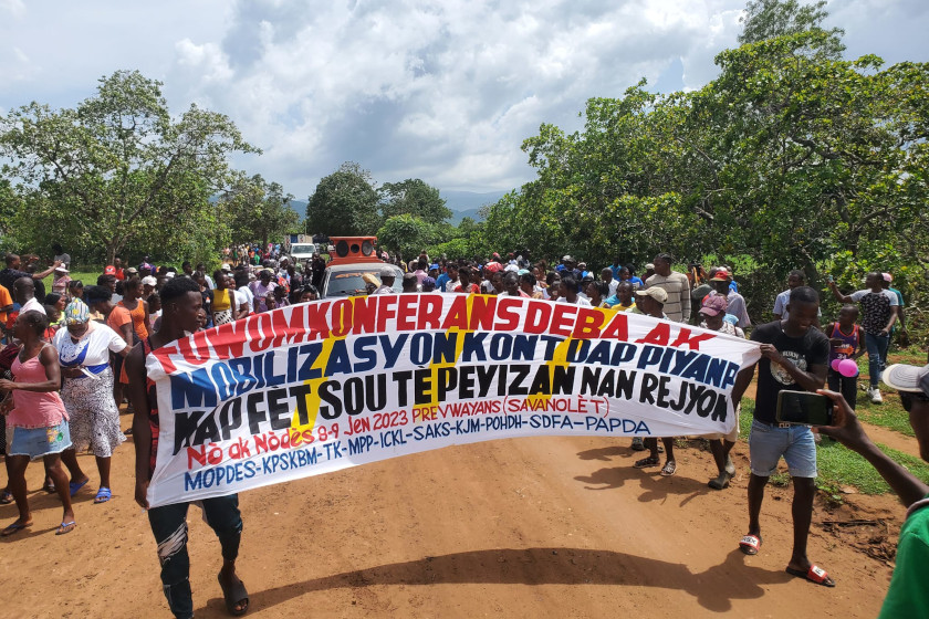 Demonstranten auf einer unbefestigten Straße; im Vordergrund tragen zwei Männer ein Banner mit einem Text auf haitianischem Kreolisch.